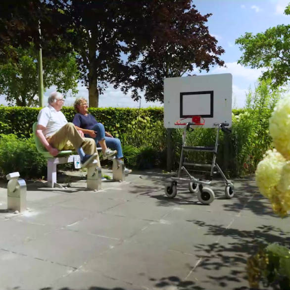 Twee ouderen zitten op een bankje op een plein. Er is een lage basketbalring waar een rollator voor staat.